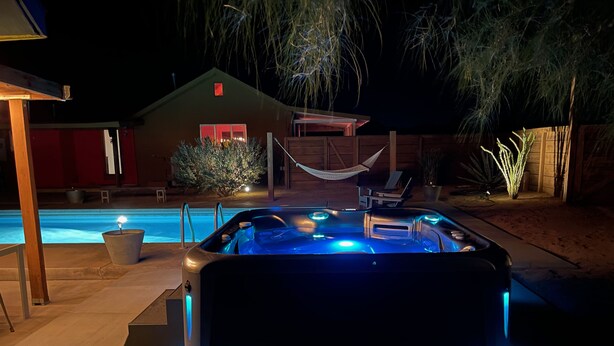Un jacuzzi con iluminación azul, una piscina, una hamaca y una casa al fondo.
