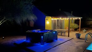 Un jacuzzi al aire libre con iluminación azul, una mesa de patio, sillas y una zona de estar cubierta con luces colgantes.