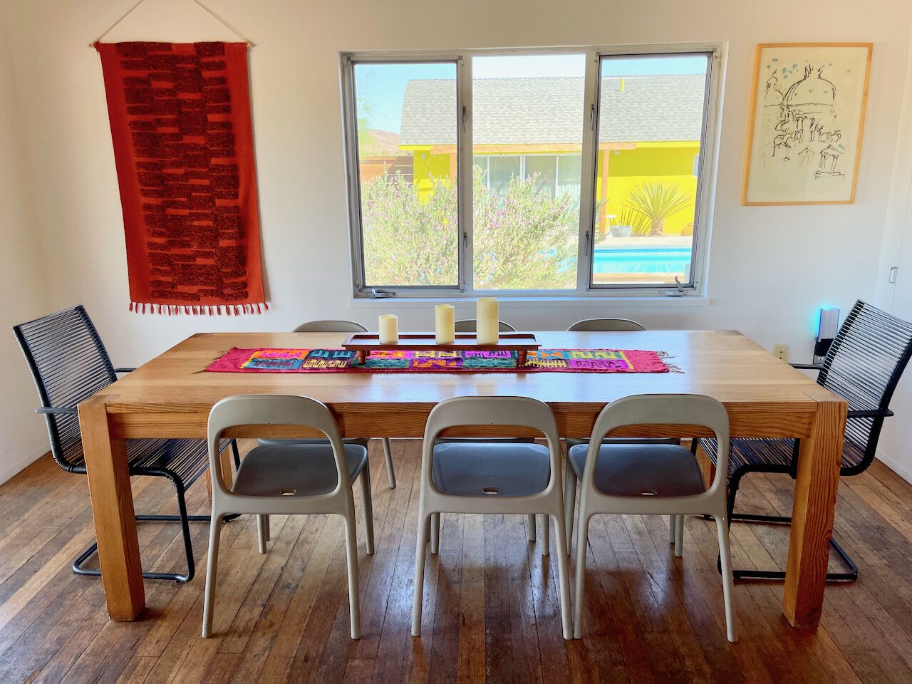 A dining area with a wooden table, four chairs, a colorful placemat, and a red tapestry.