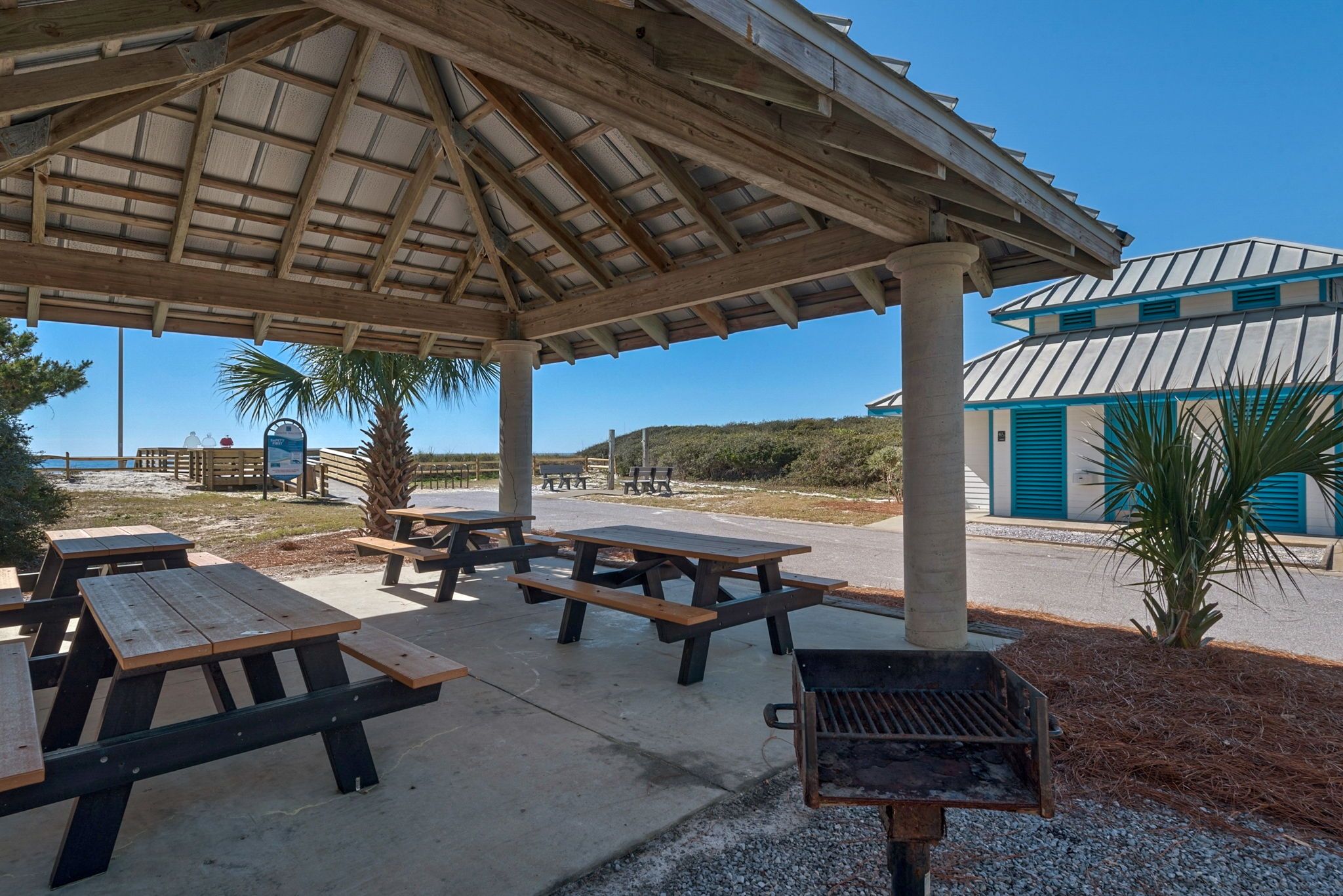 A covered picnic area with benches and a grill, a beach in the background, and a building with a blue and white facade.