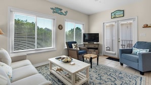 A living room with a sofa, a blue armchair, a coffee table, and a TV. There are two windows with blinds, a wall clock, and a beach-themed sign. - Breakaway~South of 30A~Short walk to the beach, Restaurants and More! (Santa Rosa Beach)