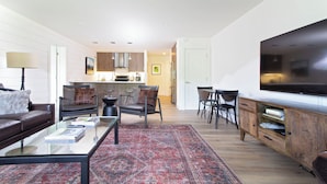 Uma sala de estar moderna com sofá, mesa de centro e poltronas, cozinha ao fundo e televisão na parede.