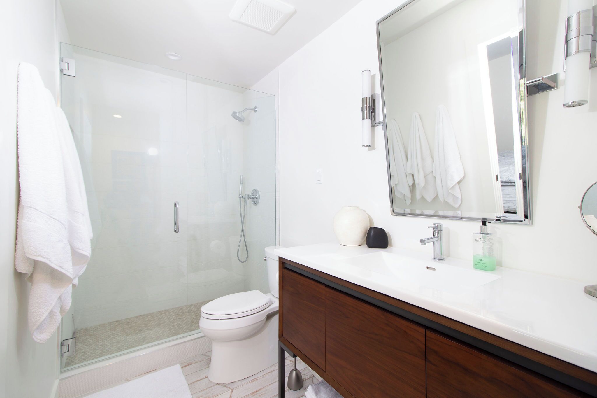 Un baño moderno con ducha envuelta en vidrio, inodoro blanco, tocador con espejo y lavamanos.