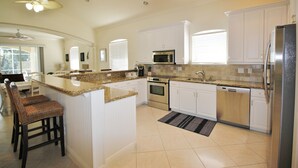 A kitchen with white cabinets, a granite countertop, stainless steel appliances, and a bar area with wicker chairs. - Large Private Pool Home-Walk/Bike to Beach & Mercato Shops & Dining (Naples)