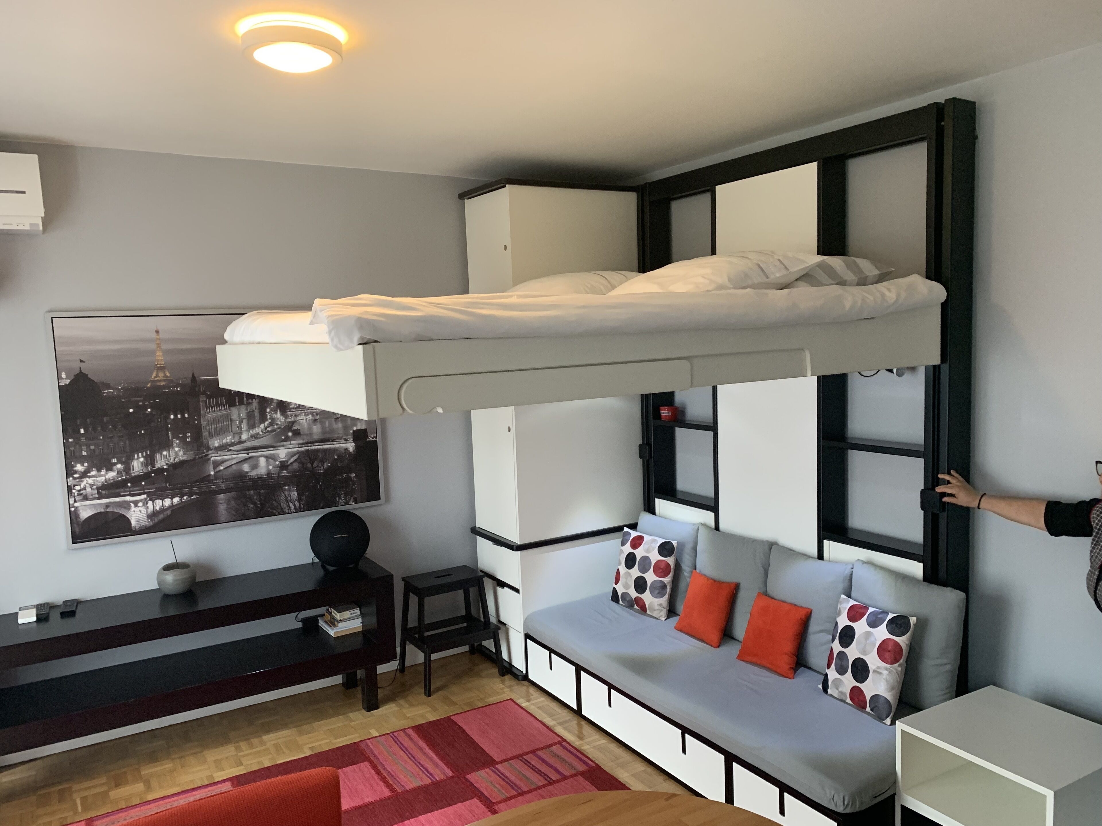 Une pièce compacte comprenant un lit superposé, un canapé agrémenté de coussins colorés, un petit bureau et un téléviseur fixé au mur qui diffuse des images d’un paysage urbain.