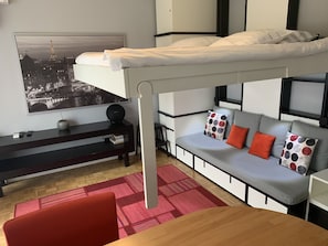 Une chambre moderne avec un lit superposé, un canapé agrémenté de coussins colorés, une petite table, un téléviseur et un tableau représentant un paysage urbain.