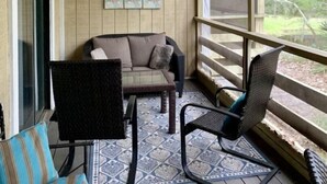 Une véranda couverte avec un ventilateur de plafond, un canapé rembourré, un fauteuil et une balustrade donnant sur un jardin.