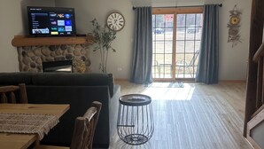 Ein Wohnzimmer mit Kamin aus Stein, einem Fernseher, einem Esstisch mit Stühlen und einem großen Fenster mit Vorhängen.