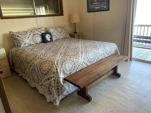 Un dormitorio con una colcha estampada, un banco de madera, una mesita de noche con una lámpara y una ventana con persianas.