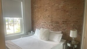 Ein Hotelzimmer mit Backsteinwand, großem Fenster, einem Bett mit weißer Bettwäsche und einem Nachttisch mit Lampe.
