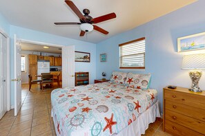 A bedroom with a bed, a nightstand, a dresser, a ceiling fan, a window with blinds, and a view of a kitchen and dining area. - Boat Parking, In Town, Pet Friendly, Golf Cart Access (Port Aransas)