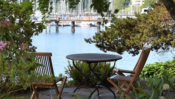 A wooden table and two chairs on a patio with a view of a water body and boats in the background.