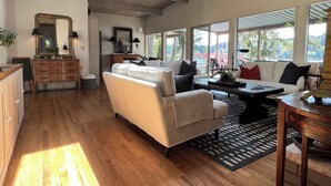 A spacious living area with a wooden ceiling, a white sofa, a coffee table, and a view of a garden through large windows.