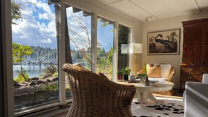 A room with large windows overlooking a body of water, a wicker chair, a white sofa, a cowhide rug, and a wooden cabinet.