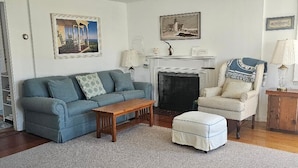 A living room with a sofa, armchair, ottoman, wooden coffee table, fireplace, and framed pictures on the wall.