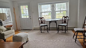 A living room with a sofa, ottoman, table, and chairs. There are two windows with a view of the outside.