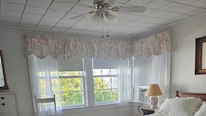 A bedroom with a wooden bed, a white dresser, a chair, a ceiling fan, and a window with curtains.