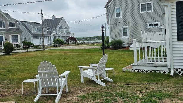 A grassy yard with two white Adirondack chairs, a white picket fence, and a house with a porch.