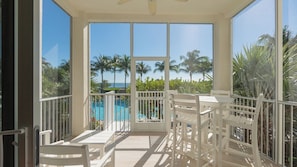 A sunlit balcony with white furniture, a pool, and palm trees in the background.