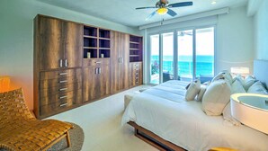 A bedroom with a large wooden wardrobe, a bed with white bedding, a ceiling fan, and a view of the ocean through large sliding doors.