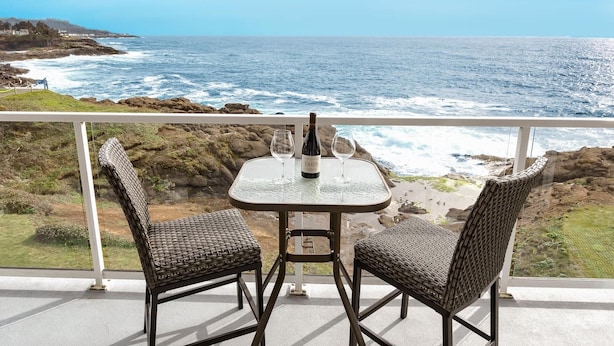A balcony with a table and two wicker chairs, overlooking a rocky coastline and the ocean.