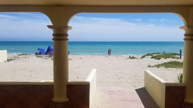 A beach view from a covered area with columns, a tent, and a few people on the sand.