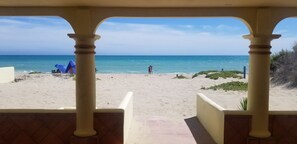 Une vue sur la plage depuis un espace abrité, doté de colonnes, avec une tente et quelques personnes sur le sable.