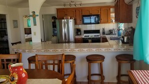 A kitchen with wooden cabinets, a stainless steel refrigerator, a microwave, a stove, and a sink. There are two wooden stools and a table with a red pitcher.