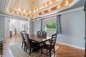 Ein Esszimmer mit einem großen Holztisch, sechs Stühlen und einer Chandelier. Eine Treppe führt zu einer anderen Ebene, und ein Fenster ist mit gemusterten Vorhängen versehen.