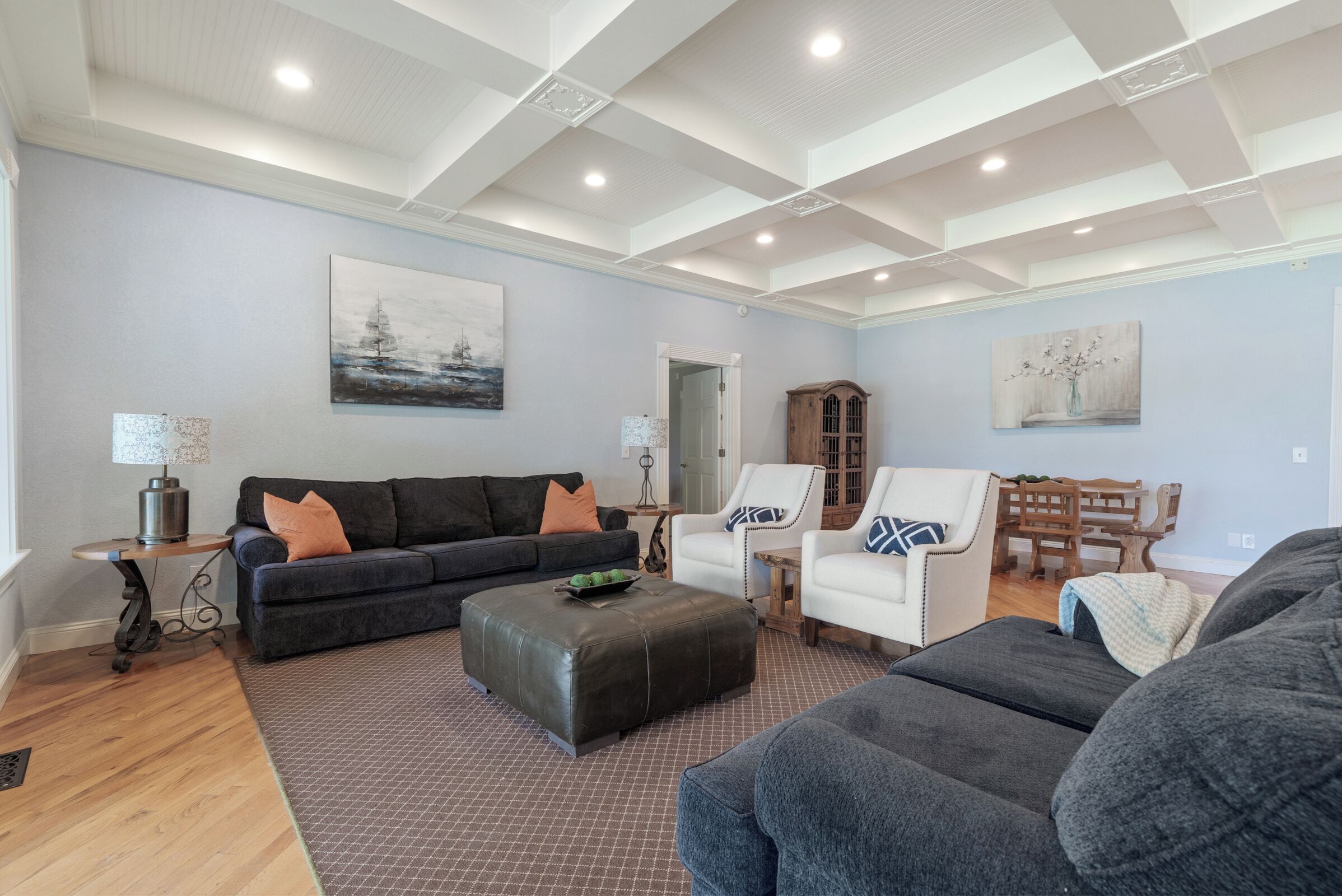 A spacious living room with a high ceiling, hardwood flooring, a black leather ottoman, a dark gray sofa, a white armchair, and a wooden dining table with chairs.