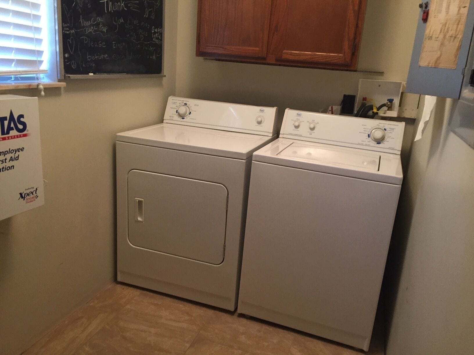 A laundry room with two stacked front-loading washers and dryers, a blackboard with writing, and a sign for employee first aid.