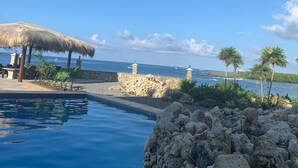 A poolside area with a clear view of the ocean, a thatched umbrella, and a rocky shoreline. -  Location with spectacular views and six minute walk to the beach!!!  (Roatan)