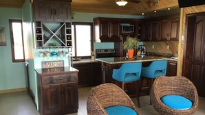 A kitchen with dark wooden cabinets, a central island with a sink, two blue bar stools, and a wicker chair with a blue cushion. -  Location with spectacular views and six minute walk to the beach!!!  (Roatan)