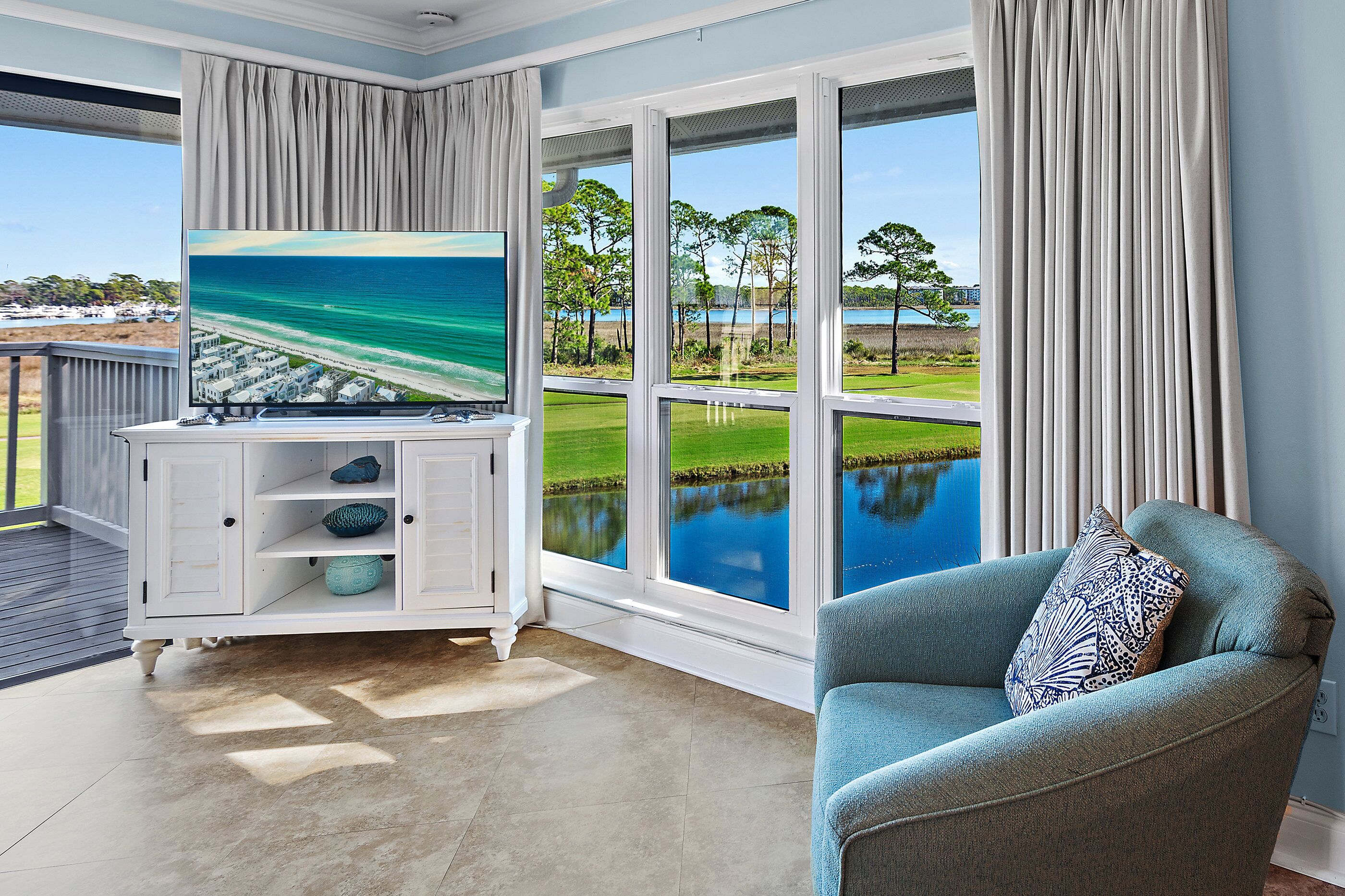 Una habitación con un ventanal grande que da a una masa de agua y vegetación, un televisor de pantalla plana sobre un mueble blanco y un sillón azul con un cojín estampado.