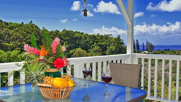 Una mesa redonda de vidrio con una cesta de mimbre que contiene frutas y flores, dos copas de vino, y una silla de mimbre en un balcón con barandilla blanca y vistas al océano y árboles.