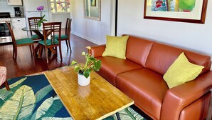 Una sala de estar moderna con un sofá de cuero marrón, cojines verdes, una mesa de centro de madera con una planta y una alfombra con motivos tropicales.