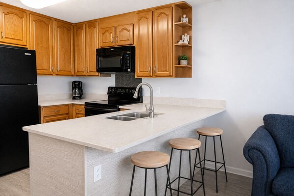 Cocina moderna con gabinetes de madera, un refrigerador negro, un microondas, una estufa y un lavamanos. Hay tres taburetes en la isla.