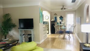 Een woonkamer met een televisie, een heldergroene bank, een eethoek met een tafel en stoelen, en een gang die leidt naar een slaapkamer.