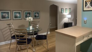 Een eethoek met een glazen tafel, vier stoelen, een plafondventilator en bloemkunst in lijsten aan de muur.