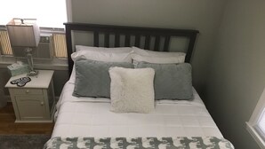 Een net opgemaakt bed met een gebloemd dekbed, twee kussens en een zacht kussen. Een nachtkastje met een lamp en zakdoekjes is zichtbaar.