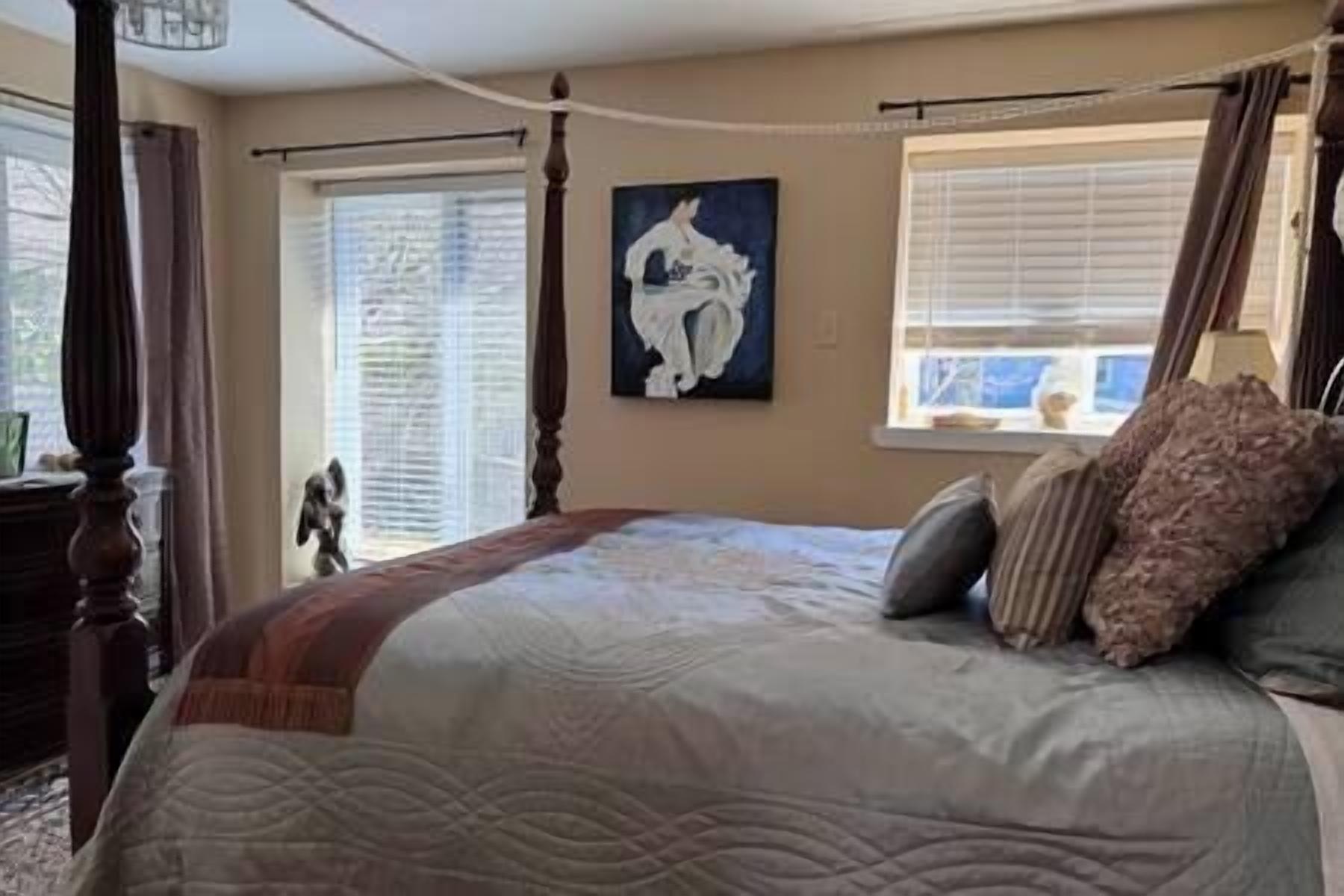 A bedroom with a large bed, a painting on the wall, and a window with blinds.