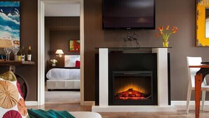 A modern living room with a fireplace, a television mounted above it, and a bed in the adjacent room. - Cottage with Puget Sound, Mount Rainier, & Olympic Views (Burien)