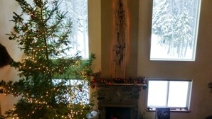 A cozy living room with a decorated Christmas tree, a stone fireplace, and large windows offering a view of snow-covered trees. - King's Cabin, just 2 miles from the Park entrance in West Glacier! (West Glacier)
