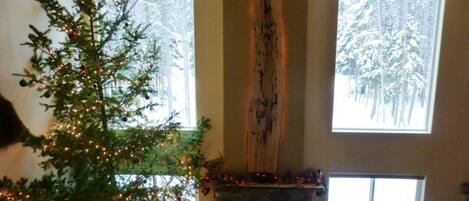 A cozy living room with a decorated Christmas tree, a stone fireplace, and large windows offering a view of snow-covered trees.