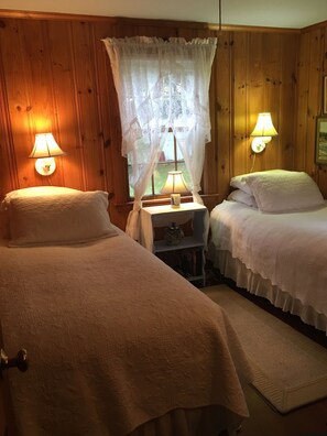 Two single beds with white comforters and pillows, set against a backdrop of wooden paneling and sheer curtains.