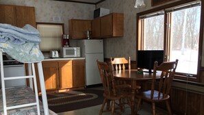 A compact kitchen with wooden cabinets, a microwave, and a refrigerator. There is a dining table with chairs and a TV on a stand. - Schuster's Fox River Resort on the Fox -Perch Place (Omro)