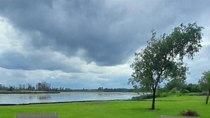 A wooden deck with two chairs and a small table, overlooking a grassy area and a body of water under a cloudy sky. - Schuster's Fox River Resort on the Fox -Perch Place (Omro)