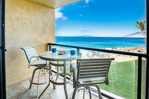 A balcony with a table and chairs, overlooking a beach with clear blue water and a distant mountain.