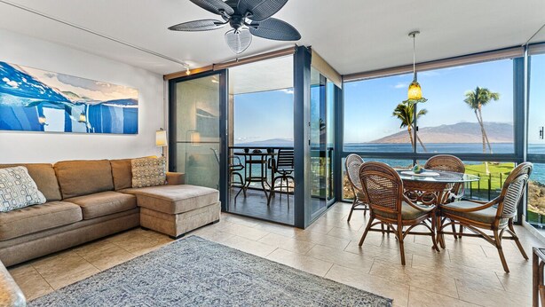 A modern living room with a sofa, a coffee table, and a dining area with chairs. There is a large window offering a view of the ocean and palm trees. - 203 - Gorgeous Remodeled Oceanfront unit with A/C! (Kihei)