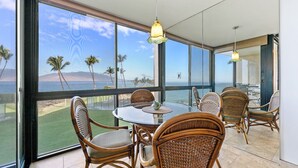 A dining area with wicker chairs and a glass table, overlooking a beach with palm trees and a view of the ocean. - 203 - Gorgeous Remodeled Oceanfront unit with A/C! (Kihei)